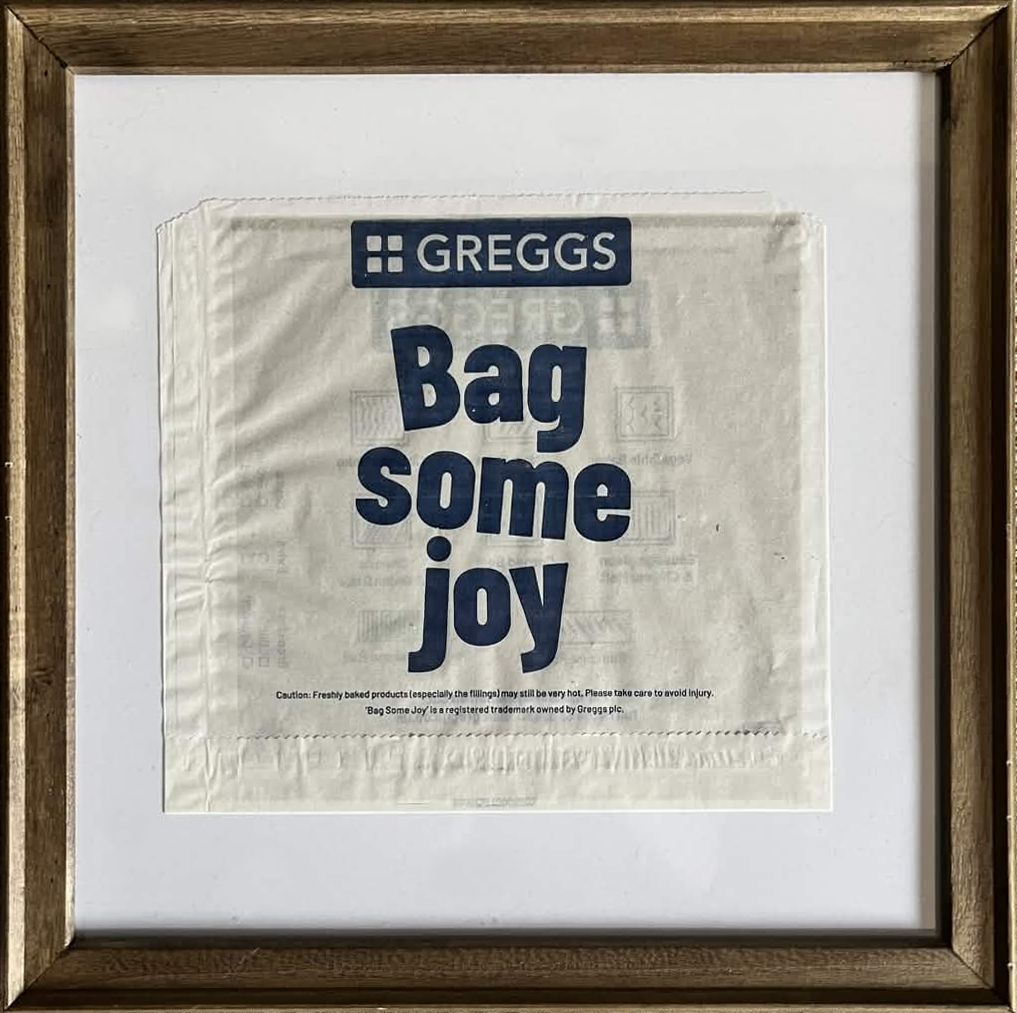 A framed square 'Bag some Joy' Greggs bag